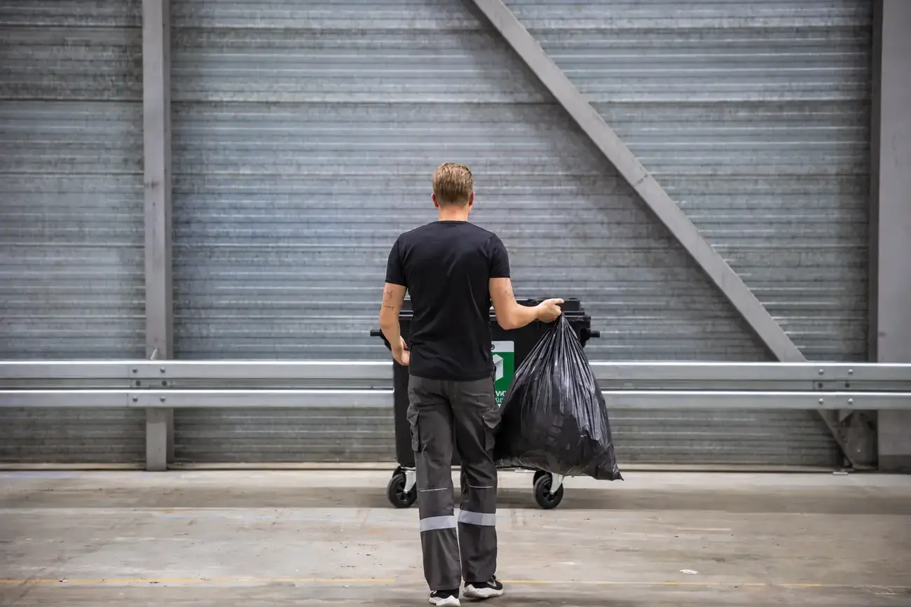 Een medewerker die een zak met restafval weggooit in de rolcontainer.