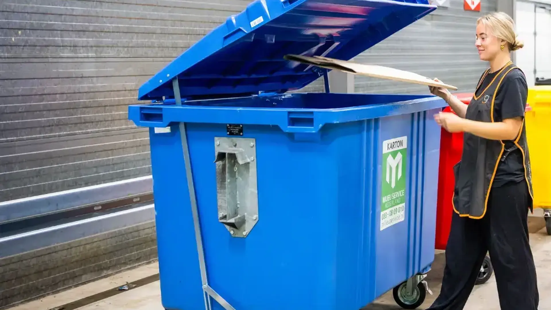 Grote papier- en kartoncontainer met voetpedaal die openstaat, waarin een platgemaakte kartonnen doos wordt gegooid. Aan de wand hangen borden met de bijbehorende afvalstroom.